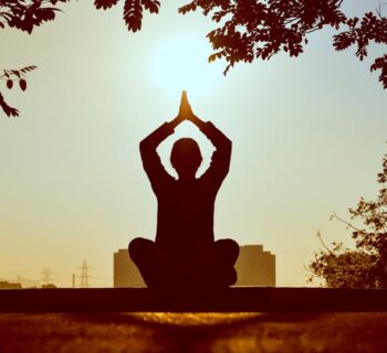 A person sitting on the ground with their palms touching over their head at sunset.